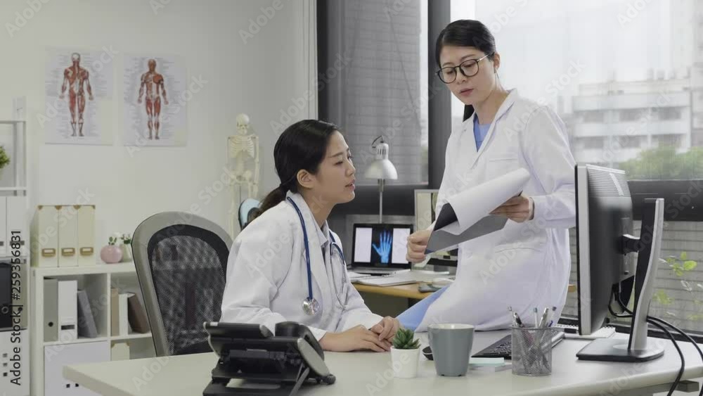 asian female medical team discussing over desktop computer monitor in working office at hospital. young woman coworker sitting on desk relying holding document pointing on screen while talking. asian female medical team discussing over desktop computer monitor in working office at hospital. young woman coworker sitting on desk relying holding document pointing on screen while talking.