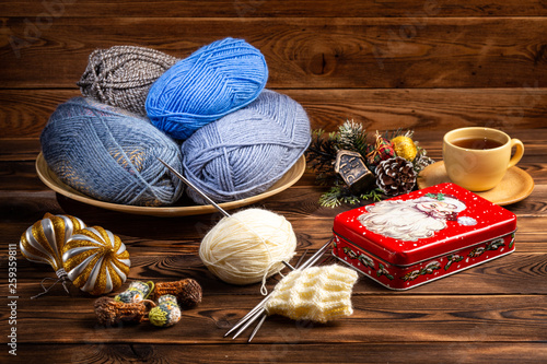 balls of gray and blue threads in a wooden plate, Christmas decorations, cup of tea and a metal box with a picture of Santa Claus knitting needles with a knitting on a wooden background