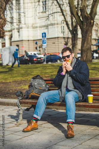 young smiling man eating burger and drink coffee sitting on city bench