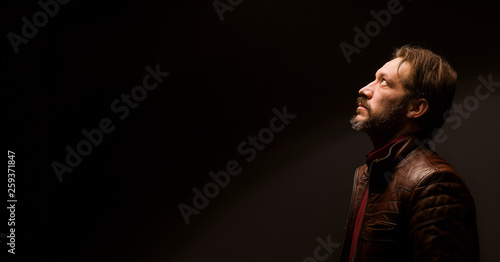 Model tests men on a black background. An adult male in a brown jacket and red shirt is looking up. Confidence and calm