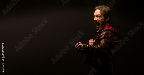 A man on a black background in a brown jacket and red shirt. With a cry goes to battle, showing aggression and expressiveness. Place for text