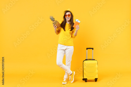 Cheerful traveler tourist woman in hat holding passport tickets, fresh pineapple fruit isolated on yellow orange background. Passenger traveling abroad on weekends getaway. Air flight journey concept.