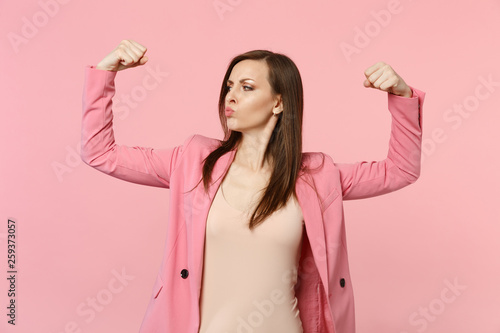 Portrait of strong funny young woman in jacket blowing lips, showing biceps, muscles isolated on pastel pink wall background in studio. People sincere emotions, lifestyle concept. Mock up copy space.