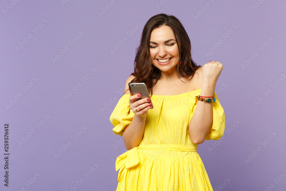 Smiling young woman in summer dress using mobile phone, typing sms message, doing winner gesture isolated on pastel violet background. People sincere emotions, lifestyle concept. Mock up copy space. Smiling young woman in summer dress using mobile phone, typing sms message, doing winner gesture isolated on pastel violet background. People sincere emotions, lifestyle concept. Mock up copy space.