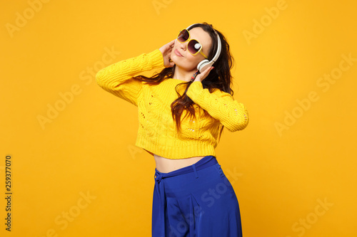 Relaxed young woman in sweater, blue trousers, sunglasses listen music putting hands on headphones isolated on yellow orange background. People sincere emotions, lifestyle concept. Mock up copy space.