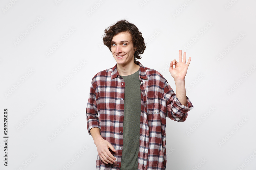 Portrait of smiling young man in casual clothes standing, looking camera showing OK gesture isolated on white wall background in studio. People sincere emotions, lifestyle concept. Mock up copy space.