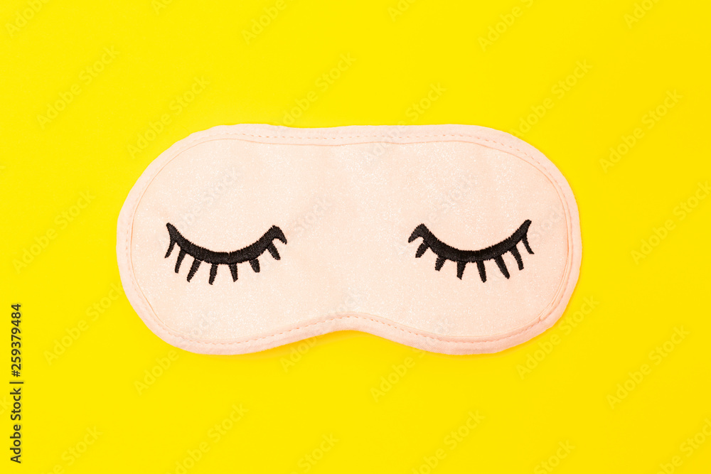 Obraz premium Pastel pink sleep mask with closed eyes embroidered on it with eyelashes on bright yellow neon paper background. Top view, flat lay. Concept of vivid dreams. Accessories for girls and young women.
