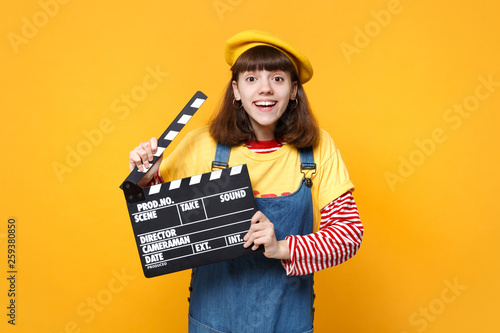 Smiling girl teenager in french beret, denim sundress holding classic black film making clapperboard isolated on yellow wall background. People sincere emotions, lifestyle concept. Mock up copy space. © ViDi Studio