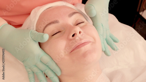 Smiling girl takes a facial massage in a beauty salon. Masseur kneads the girl's ears. Beauty and health. Close-up of female face and gloved hands