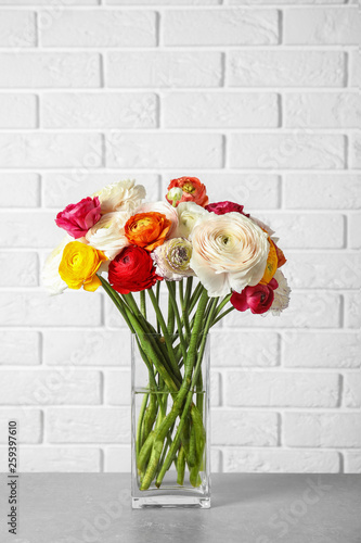 Vase with beautiful spring ranunculus flowers on table near brick wall