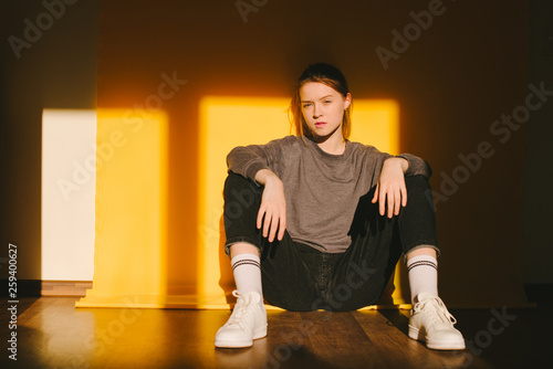 Stylish girl sits on the ground on an orange background, wear casual clothes and sneakers, looks into the camera. Portrait of a girl in a studio with sunshine.
