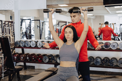 A young girl wearing sportswear in a gym performs dumbbell exercises, the coach helps her