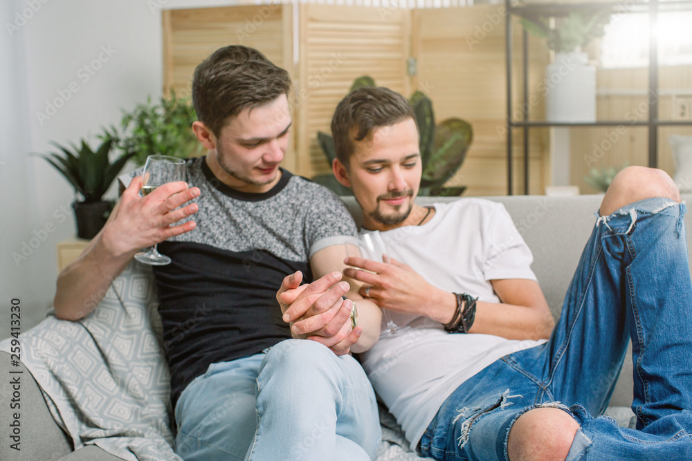 Young gay men holding hands on sofa at home. Shy gay couple showing love and romance, drinking wine Young gay men holding hands on sofa at home. Shy gay couple showing love and romance, drinking wine