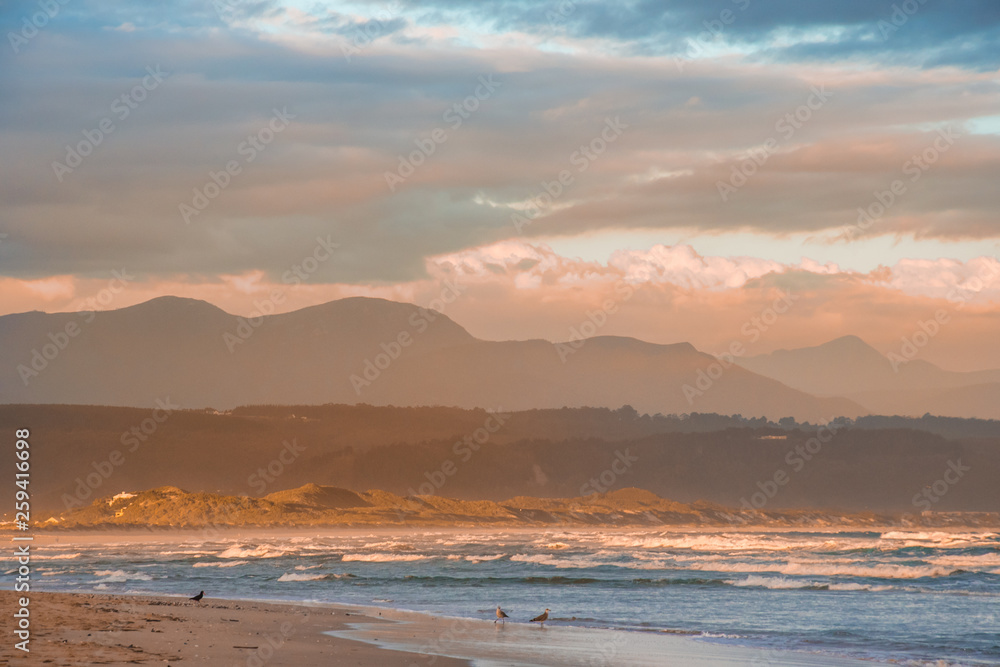Rays of golden evening light falling on the Plettenberg Bay beach at sunset, with mountains in the distance. Garden Route, Western Cape, South Africa Rays of golden evening light falling on the Plettenberg Bay beach at sunset, with mountains in the distance. Garden Route, Western Cape, South Africa