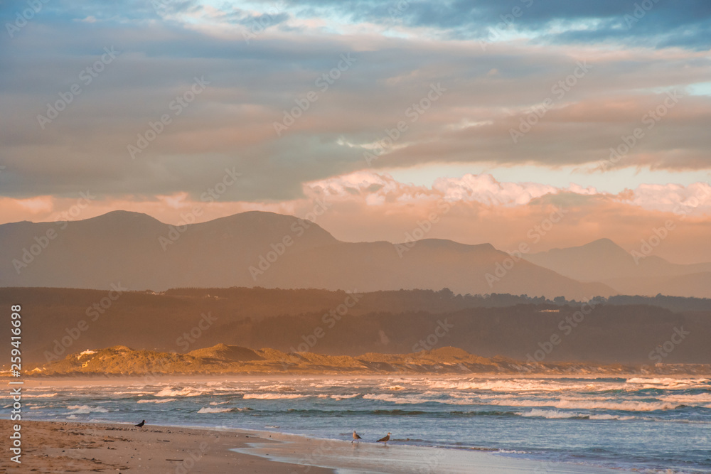 Rays of golden evening light falling on the Plettenberg Bay beach at sunset, with mountains in the distance. Garden Route, Western Cape, South Africa