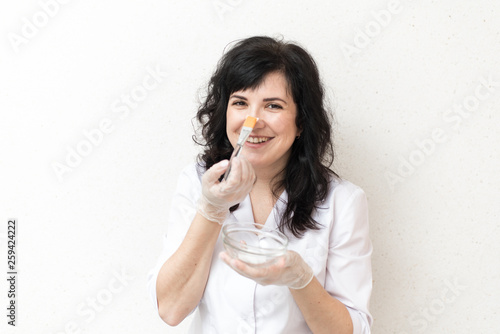 advertising campaign for cosmetology. A girl in a white medical gown mixes a mask on a white background and applies it to her face. Smile and emotions