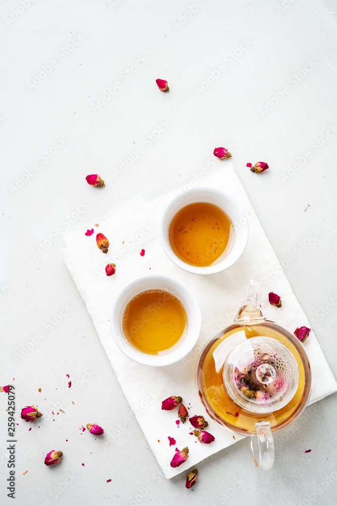 Top view of two white cups and transparent teapot with herbal tea of dried pink roses buds over textile napkin on gray background with copy space. Brewing and Drinking tea. Top view of two white cups and transparent teapot with herbal tea of dried pink roses buds over textile napkin on gray background with copy space. Brewing and Drinking tea.