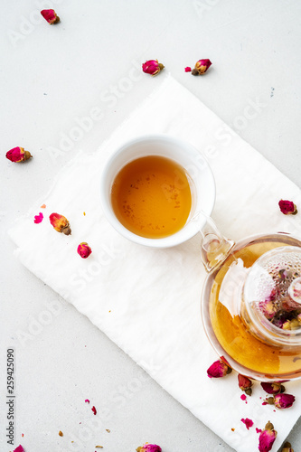 Top view of two white cups and transparent teapot with herbal tea of dried pink roses buds over textile napkin on gray background with copy space. Brewing and Drinking tea.