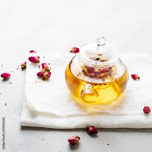 Herbal tea with dried pink flower roses buds in transparent teapot over gray background with copy space. Brewing and Drinking tea.