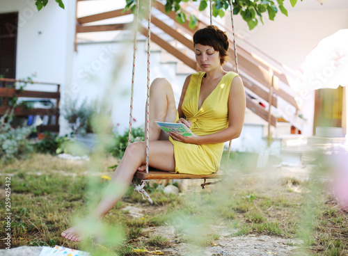 A young woman is swinging on a swing and writing postcards and letters to her friends in front of her summer house in bushes and greenery. Villa vacations. Lifestyle landscape. 