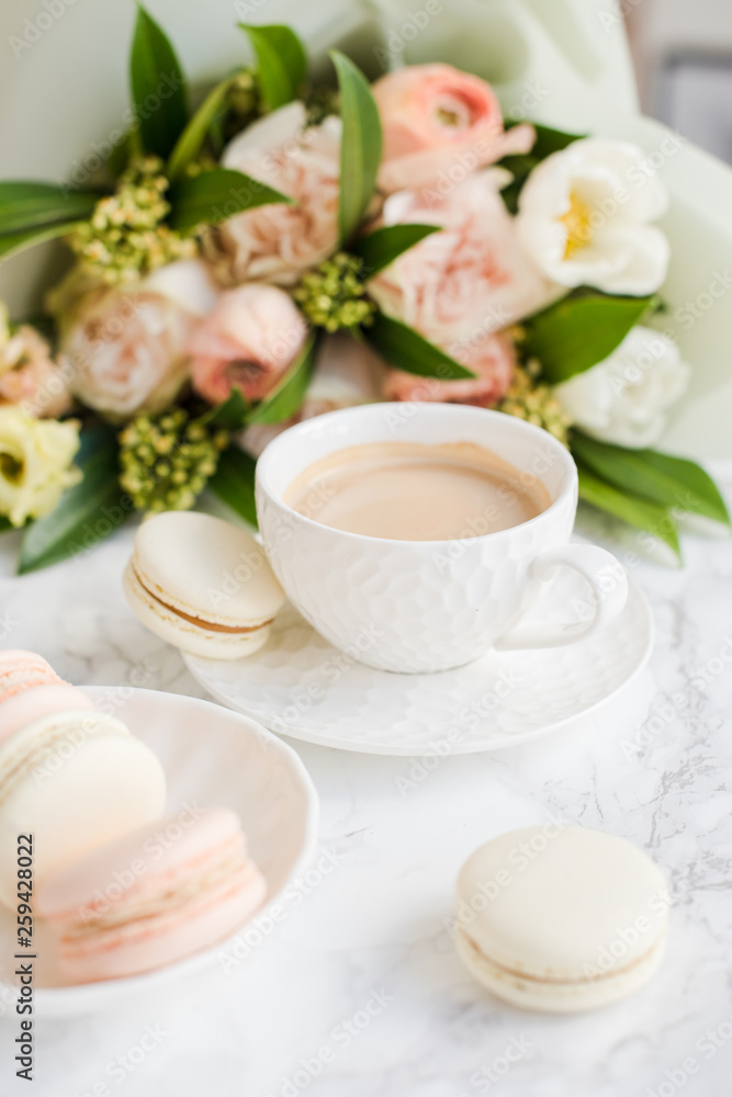 Elegant sweet dessert macarons, cup of coffee and pastel colored beige flowers bouquet on white marble Elegant sweet dessert macarons, cup of coffee and pastel colored beige flowers bouquet on white marble