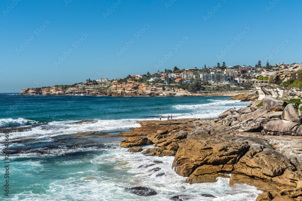 Sydney, Australia - February 11, 2019: Wide shot of Bronte beach with neighborhoods above and rocks to the north and south. Blue sea and blue sky. Waves crashing on rocks.