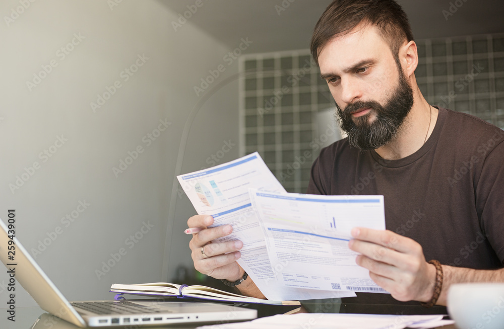 Bearded young man sitting at table looking at documents and thinking. Business man going through paperwork at home office. Bearded young man sitting at table looking at documents and thinking. Business man going through paperwork at home office.