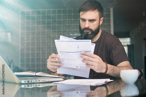 Bearded young man sitting at table looking at documents and thinking. Business man going through paperwork at home office.