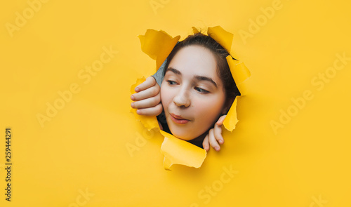 Funny teenage girl peeping through hole on yellow paper. The concept of surprise, joyful mood from what he saw. Discounts, sales, surprise. Copy space. photo