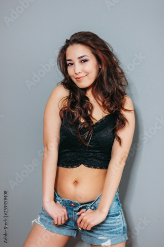 Happy sensual smiling asian young woman with dark long curly hair on grey wall background. Girl having fun at the party