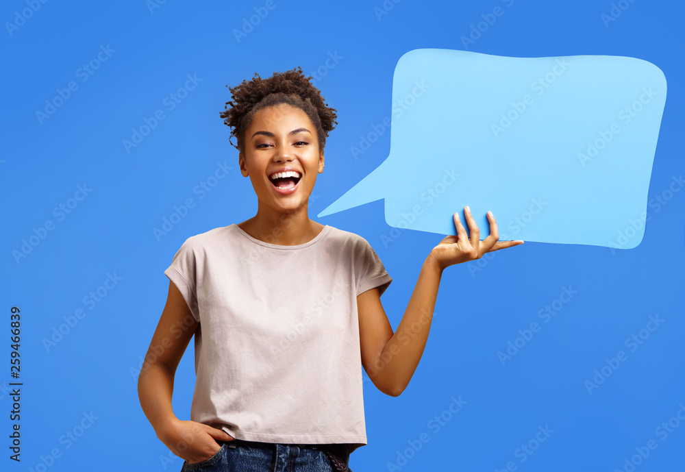 Happy laughing girl holds blue speech bubble banner. Photo of african american girl wears casual outfit on blue background. Emotions and pleasant feelings concept. - obrazy, fototapety, plakaty 