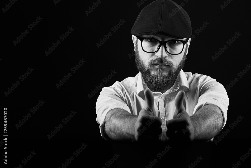 young handsome man in glasses with a beard and a mustache in a black cap and a white shirt shows a variety of hand gestures, a dark studio background