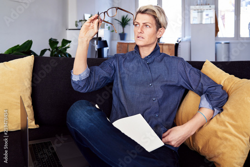 Business woman with short hair looks into glasses, makign notes, using laptop at modern apartment. Concept of young entrepreneur works on start-up. photo