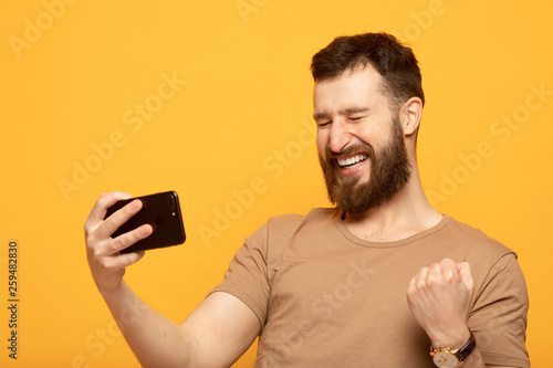 Portrait of an excited man standing over yellow background, holding mobile phone, celebrating 