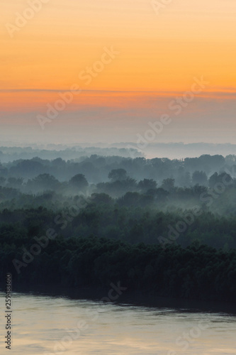Mystical view on riverbank of large island with forest under haze at early morning. Mist among layers from tree silhouettes under warm predawn sky. Morning atmospheric landscape of majestic nature.
