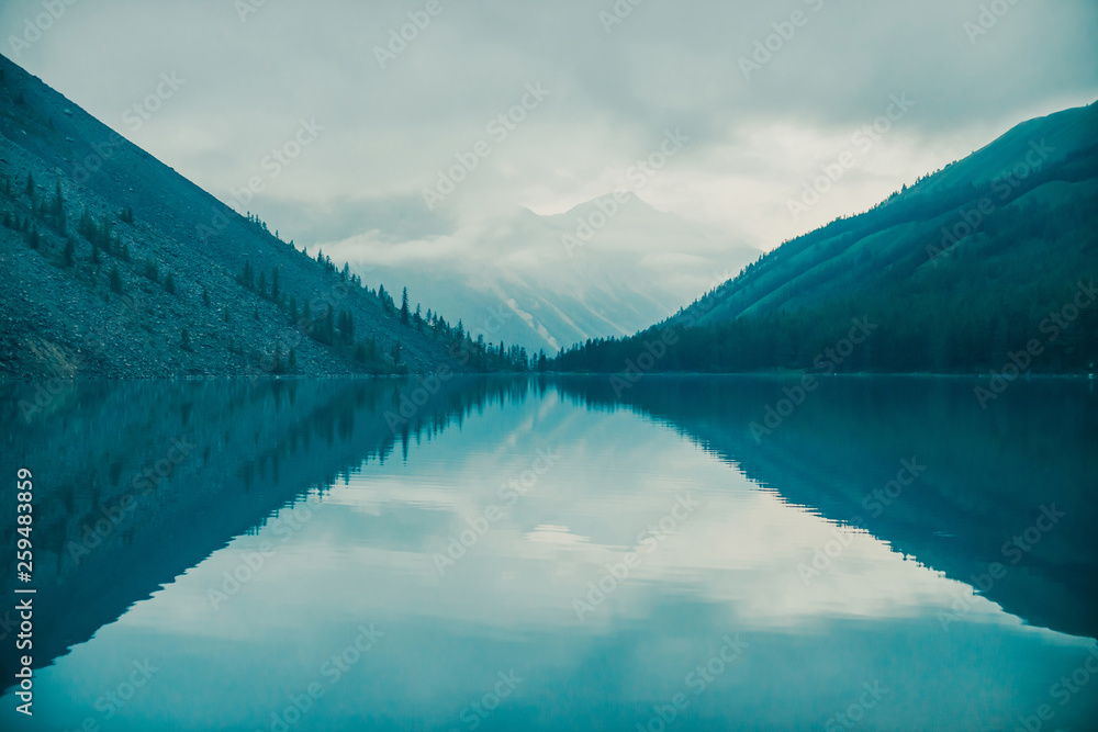 Amazing silhouettes of mountains and low clouds reflected on mountain lake. Beautiful ripples on water mirror. Cloudy sky in highlands. Atmospheric ghostly landscape. Wonderful mystic mountainscape.