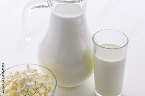 Fresh milk in a glass poured from a jug with a bowl of cottage cheese on a white background. Dairy products for breakfast are very healthy.