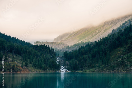 Fototapeta Naklejka Na Ścianę i Meble -  Ghostly forest near mountain lake in early morning. Mountain creek flows into lake. Ripple on smooth water surface. Low clouds. Dark calm atmospheric misty woodland landscape. Tranquil atmosphere.
