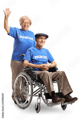 Senior volunteer woman waving and pushing a senior volunteer man in a wheelchair