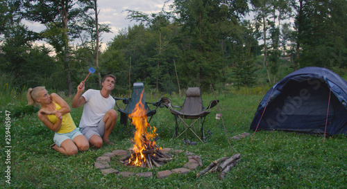 Caucasian man and woman sitting by campfire waving a fly swatter to repel bugs.