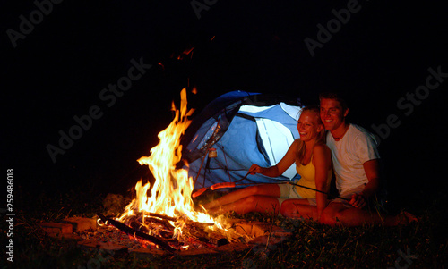 CLOSE UP: Happy couple camping in the wild roasting hot dogs by their tent.