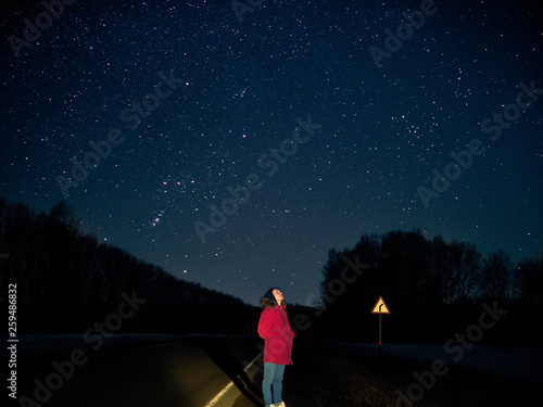 A young guy stands at night on the edge of the road in a red jacket and looks at the sky dotted with stars.