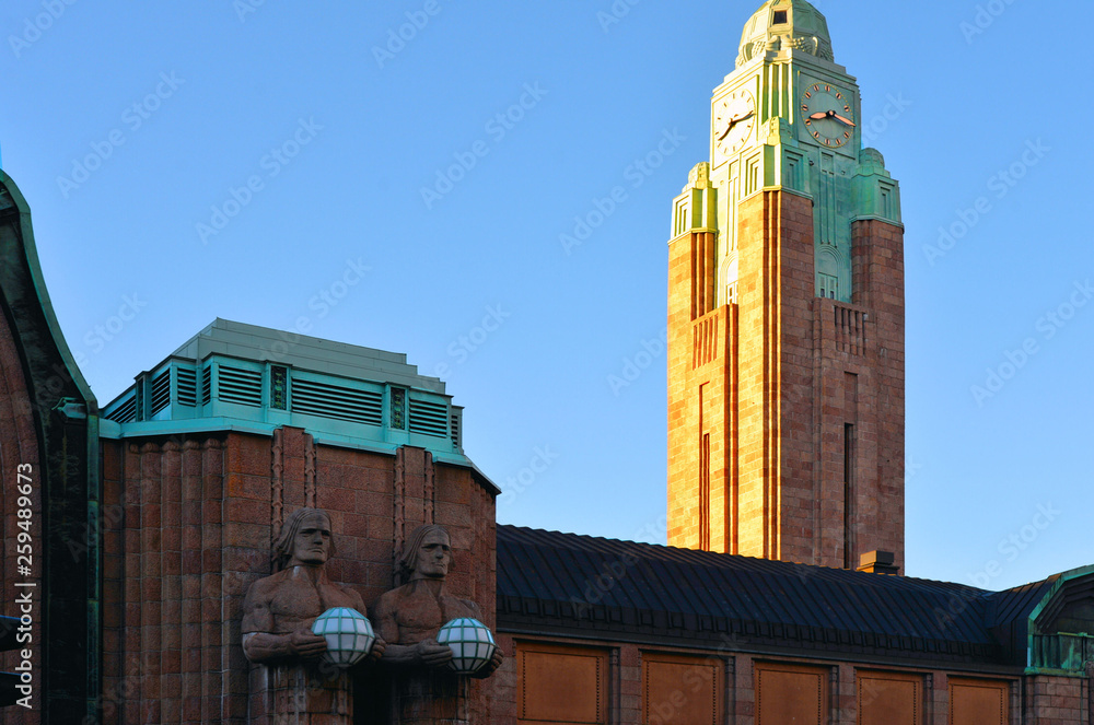 Fototapeta premium Helsinki, Finland. View Of Statues On Entrance To Helsinki Central Railway Station in shadows and sun Illuminated clock tower in back ground.