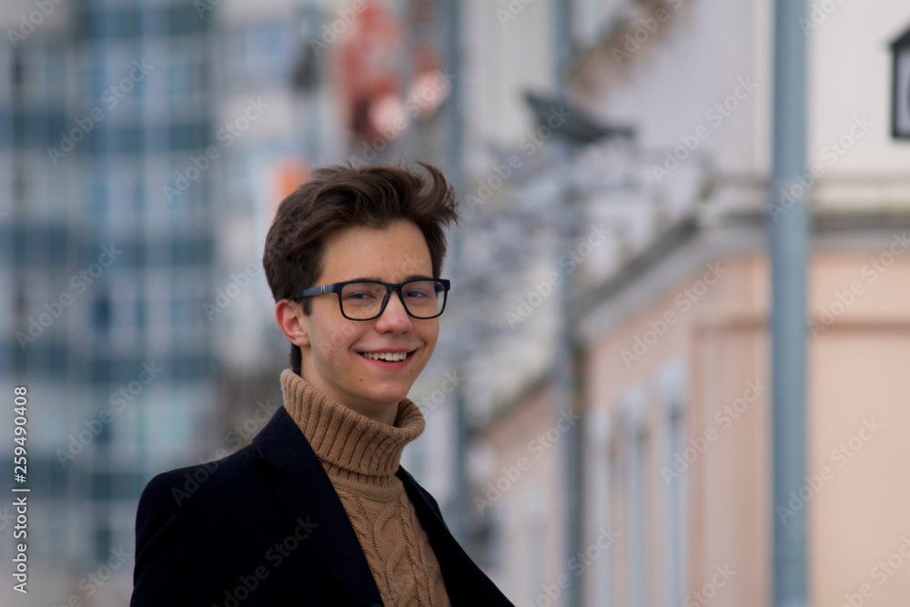 A young man posing with a smile, among the cityscape. Dressed in a black light overcoat, black-rimmed glasses. A young man posing with a smile, among the cityscape. Dressed in a black light overcoat, black-rimmed glasses.