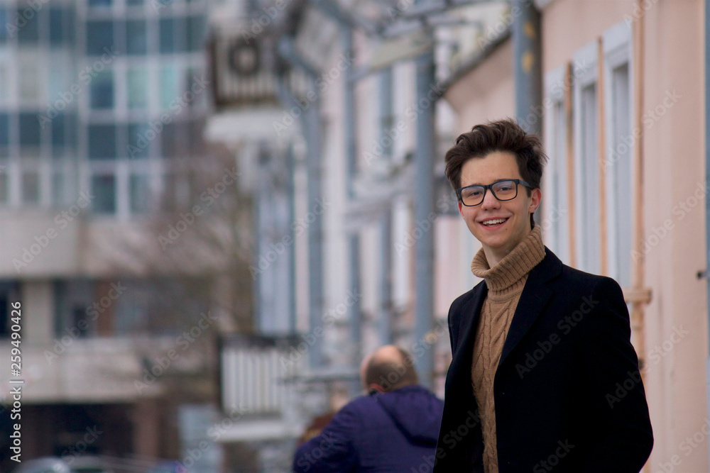 A young man posing with a smile, among the cityscape. Dressed in a black light overcoat, black-rimmed glasses. A young man posing with a smile, among the cityscape. Dressed in a black light overcoat, black-rimmed glasses.