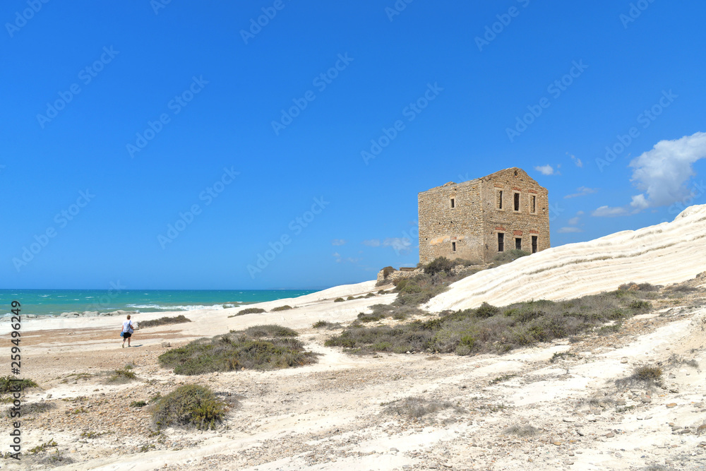 empty white beach with old ruins of abandoned stone house on the rocks and summer blue sky and sea in background in Sicily Italy