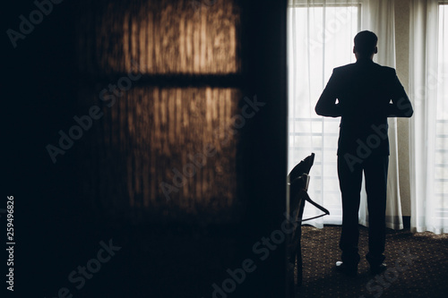 groom getting ready in the morning before wedding ceremony, putting on jacket on shirt in room silhouette.