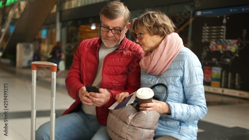 Couple of mature people consults the smartphone waiting for the train. Husband and wife waiting for the train sit on a bench at the railway station