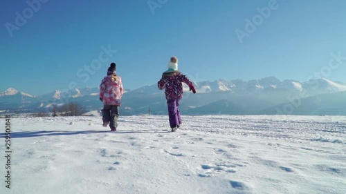 Charming kids in outwear running on snowy terrain with beautiful mountains on background in sunshine