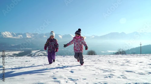 Charming kids in outwear running on snowy terrain with beautiful mountains on background in sunshine